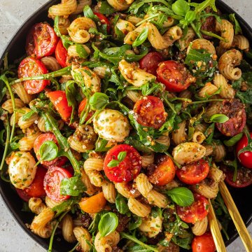 Bunter Nudelsalat mit Tomaten und Rucola auf einem großen Teller mit Deko im Bildrand.