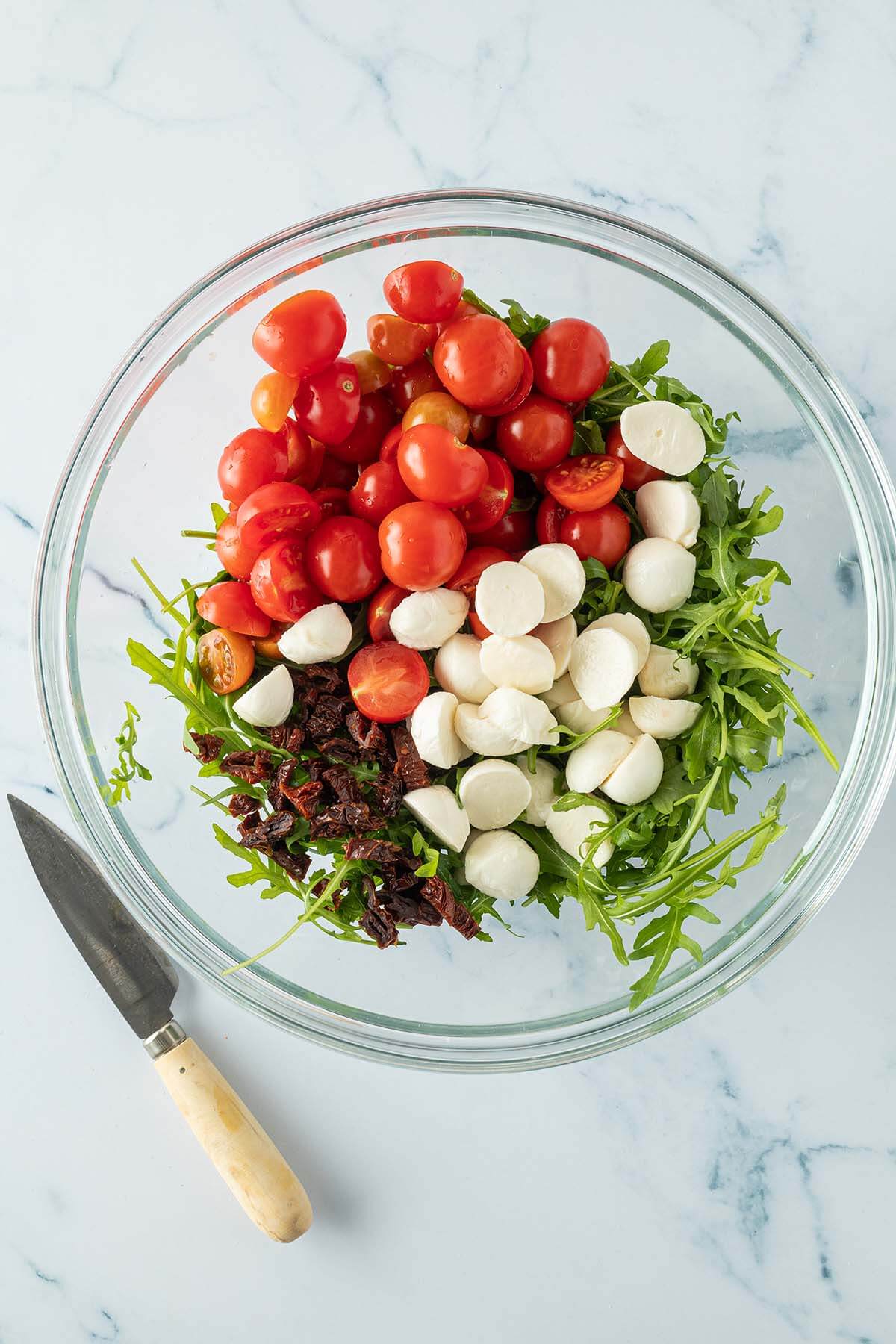 Große Glasschüssel mit Rucola, halbierten Cherry Tomaten, halbierten Mozzarella Kugeln und kleingeschnittenen, getrockneten Tomaten.