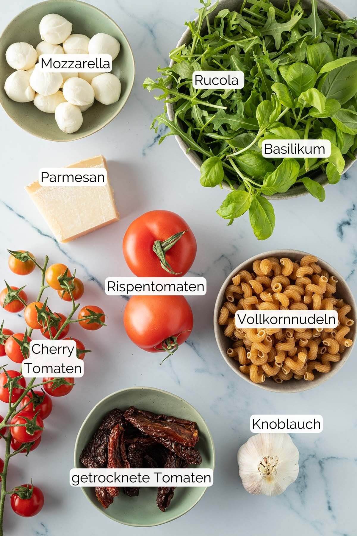 Foto aller Zutaten, die für den Nudelsalat benötigt werden, mit Beschriftungen.
