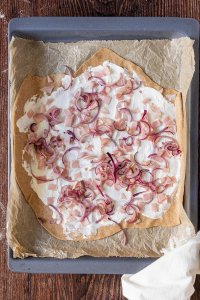 Gesunder Vollkorn Flammkuchenteig wird mit Frischk&auml;se, Schinkenw&uuml;rfel und angebratenen Zwiebelringen belegt.