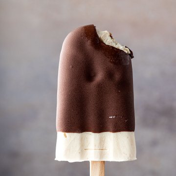 Eine Hand h&auml;lt ein angebissenes Eis am Stiel mit Schoko&uuml;berzug vor einem grauen Hintergrund.