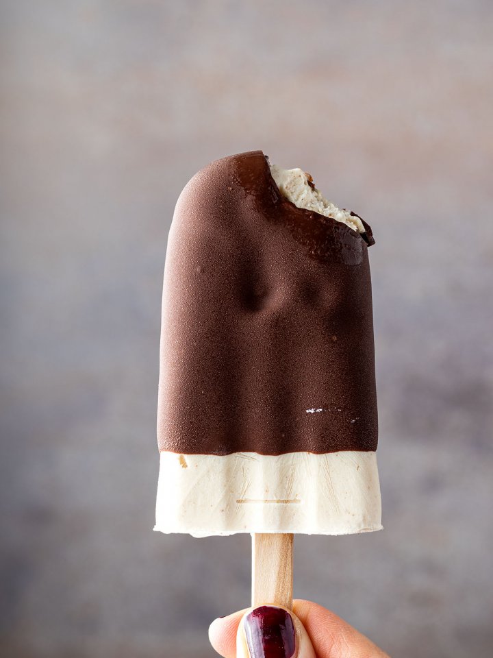Eine Hand h&auml;lt ein angebissenes Eis am Stiel mit Schoko&uuml;berzug vor einem grauen Hintergrund.