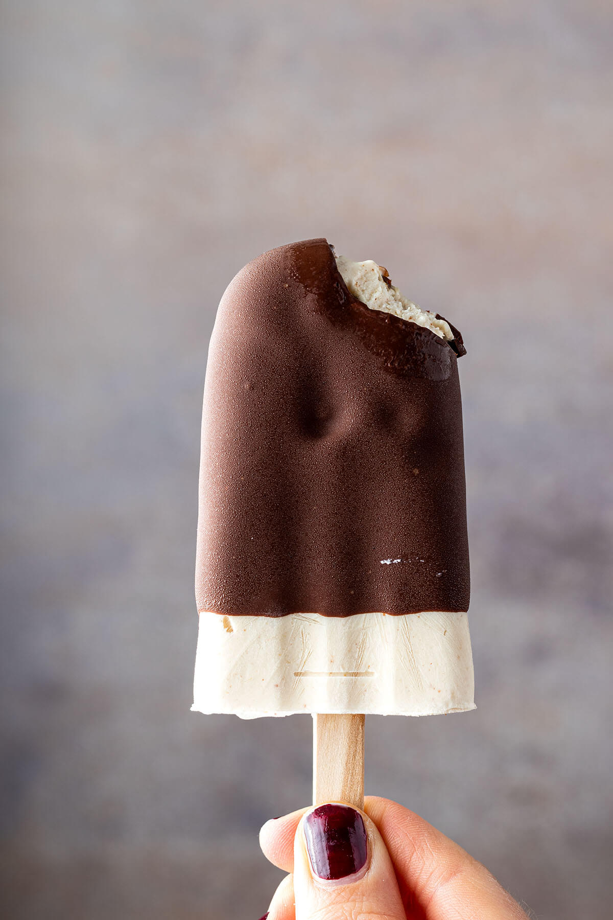 Eine Hand h&auml;lt ein angebissenes Eis am Stiel mit Schoko&uuml;berzug vor einem grauen Hintergrund.
