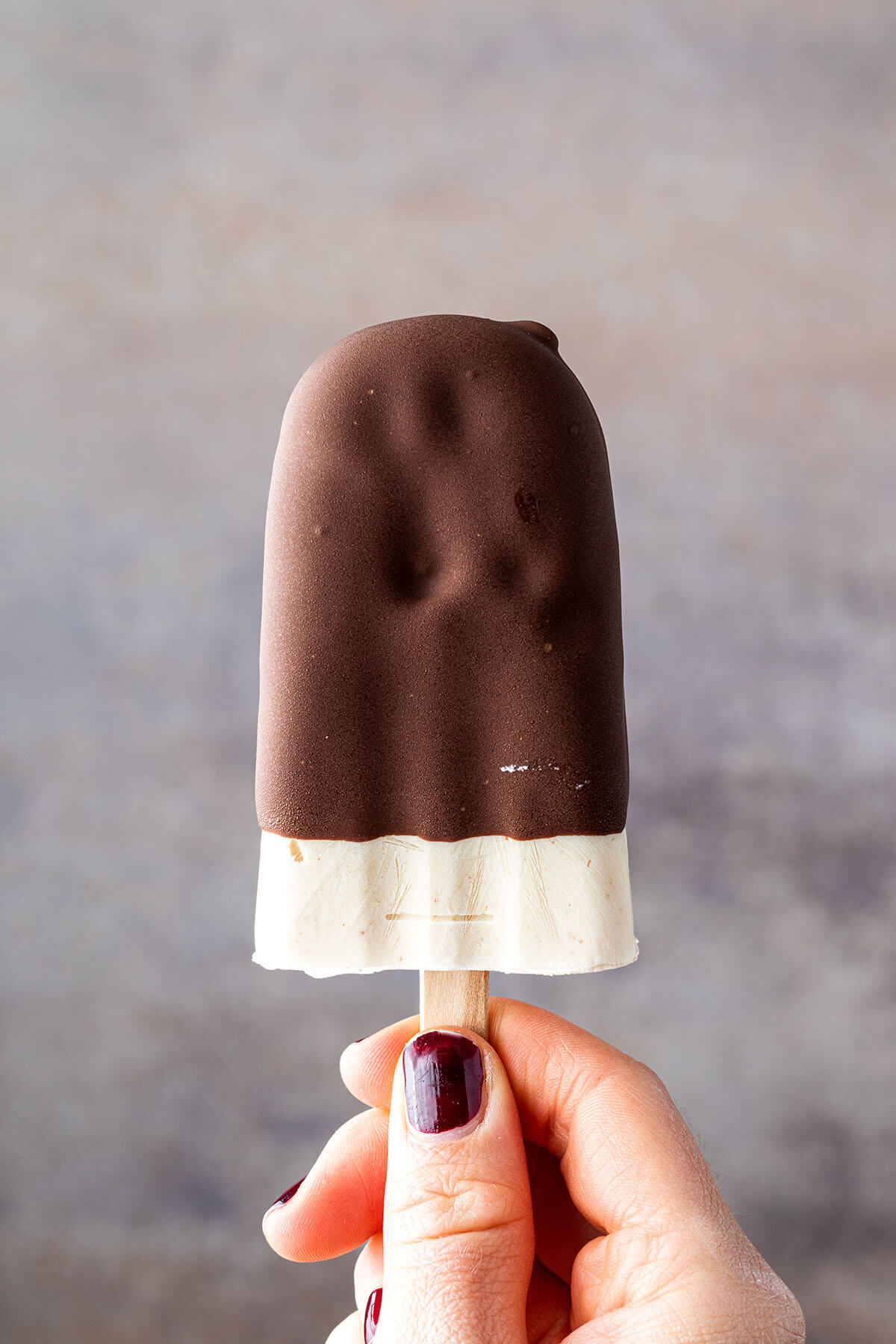 Ein Milch-Eis am Stiel mit Schoko&uuml;berzug wird von einer Hand gehalten vor einem braun-grauen Hintergrund.