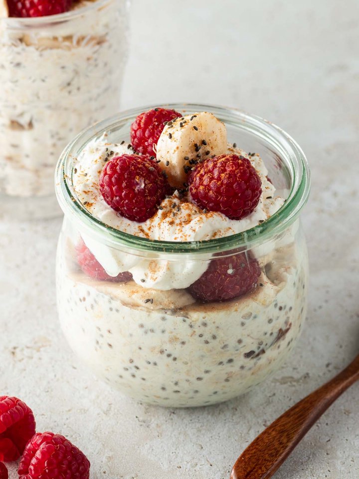 Ein gesundes Fr&uuml;hst&uuml;ck im Glas mit frischen Himbeeren und Bananenscheiben dekoriert auf einem beigen Untergrund. Im Hintergrund ein zweites Glas.