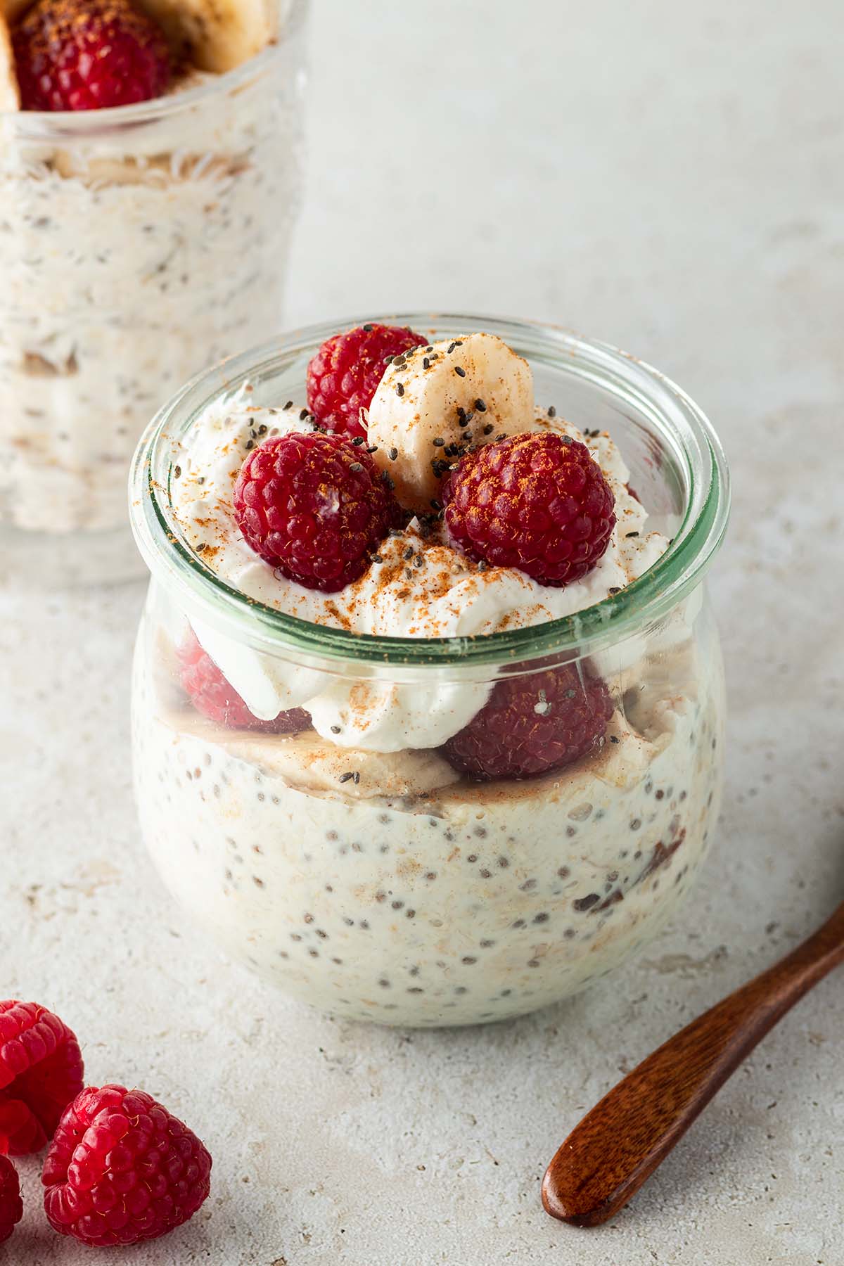 Ein gesundes Fr&uuml;hst&uuml;ck im Glas mit frischen Himbeeren und Bananenscheiben dekoriert auf einem beigen Untergrund. Im Hintergrund ein zweites Glas.
