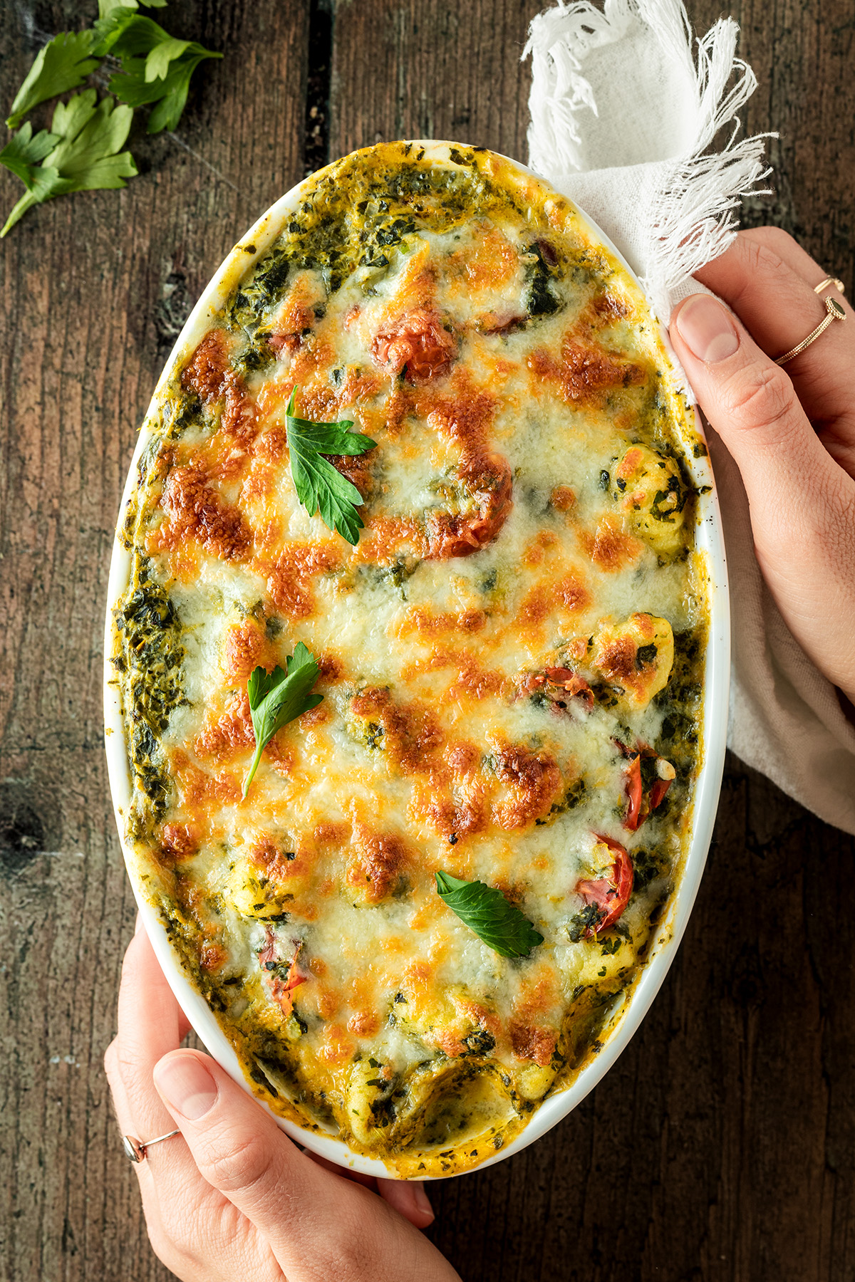 Zwei H&auml;nde halten eine Auflaufform mit einem Gnocchi Auflauf mit Spinat und viel K&auml;se.