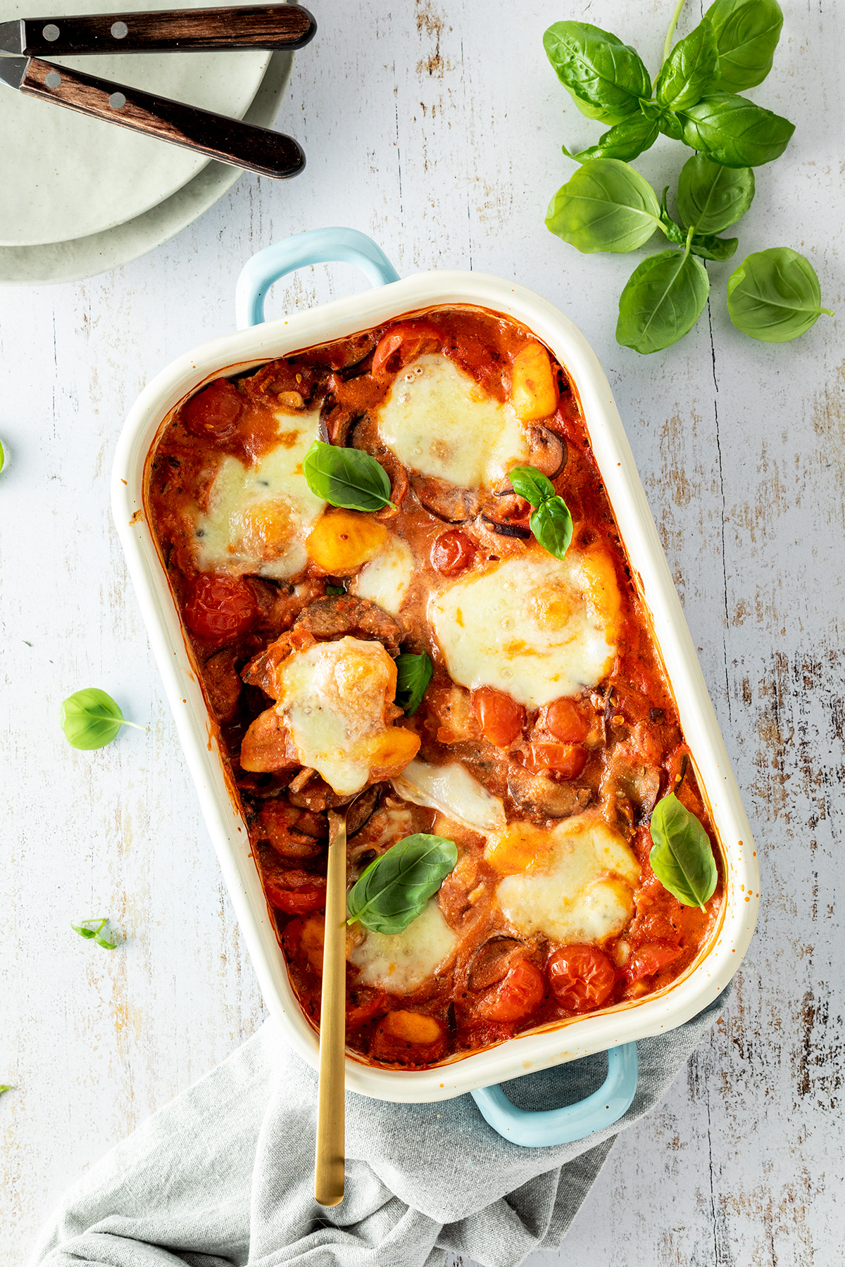 Ein vegetarischer Gnocchi Auflauf mit Tomate Mozzarella und frischem Basilikum serviert in einer Auflaufform. Daneben Teller und noch mehr Basilikum.