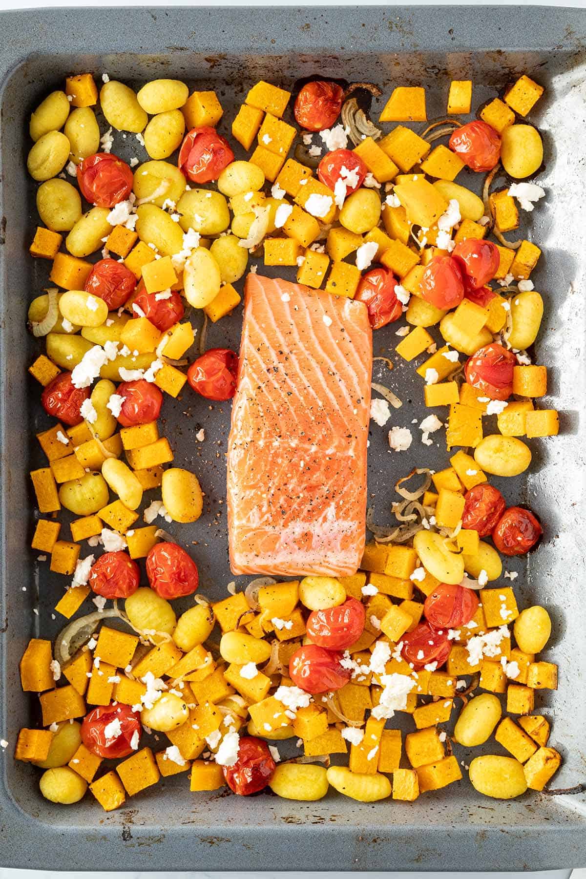 Ger&ouml;stete Gnocchi und Gem&uuml;se auf einem Blech mit einem rohen Lachsfilet in der Mitte und etwas Feta &uuml;ber dem Gem&uuml;se.