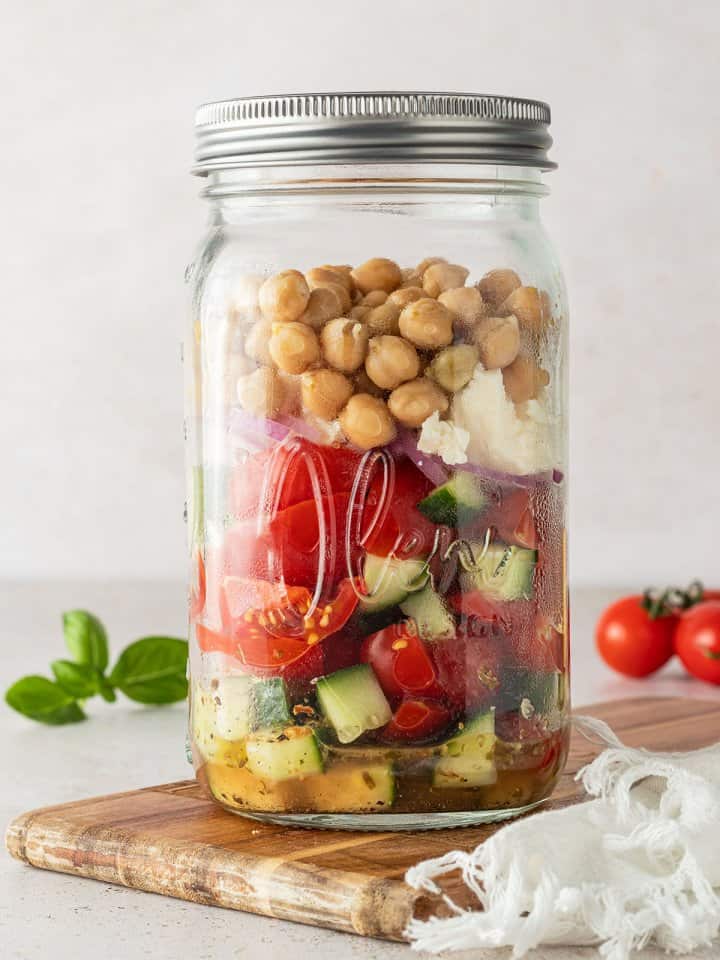 Griechischer Salat mit Kichererbsen in einem gro&szlig;en Schraubglas vor beigen Hintergrund, Deko im Bildrand.