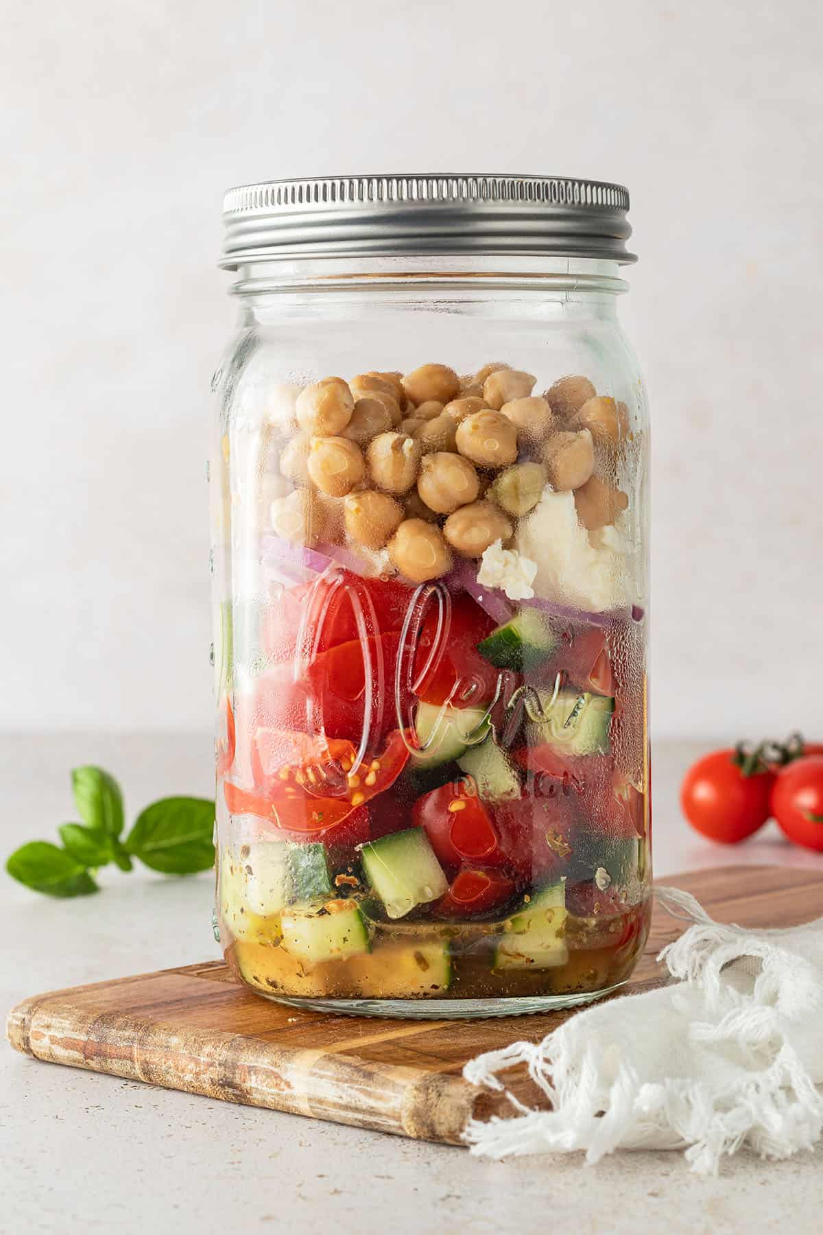 Griechischer Salat mit Kichererbsen in einem gro&szlig;en Schraubglas vor beigen Hintergrund, Deko im Bildrand.