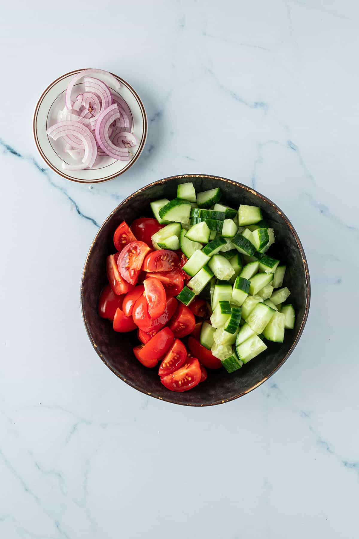 Eine Sch&uuml;ssel mit klein geschnittenen Tomaten und Gurken, daneben ein kleiner Teller mit Zwiebelringen.