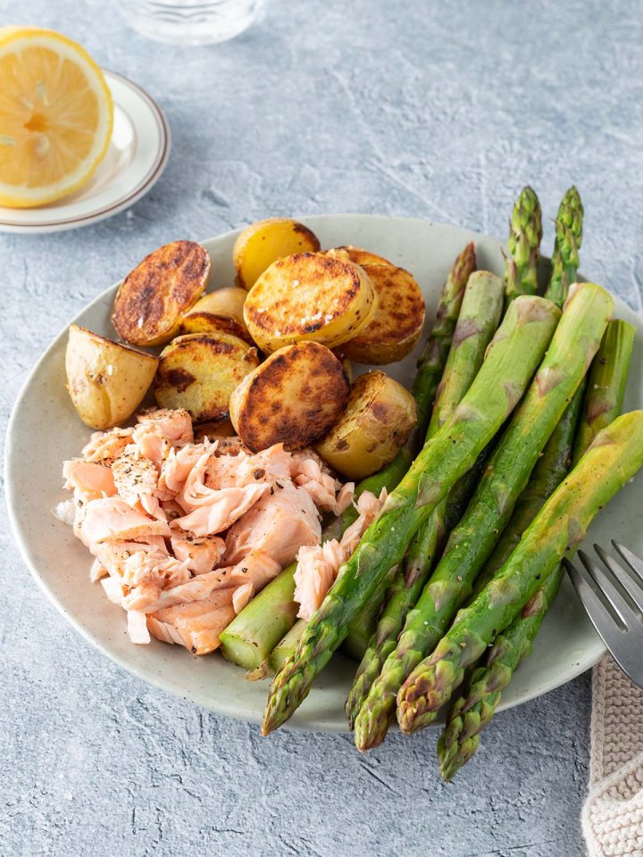 Ein Teller mit gr&uuml;nem Spargel, knusprig angebratenen Kartoffeln und ein kleine St&uuml;cke zerrissener Lachs, eine Glas Wasser und eine Zitronenh&auml;lfte im Hintergrund.
