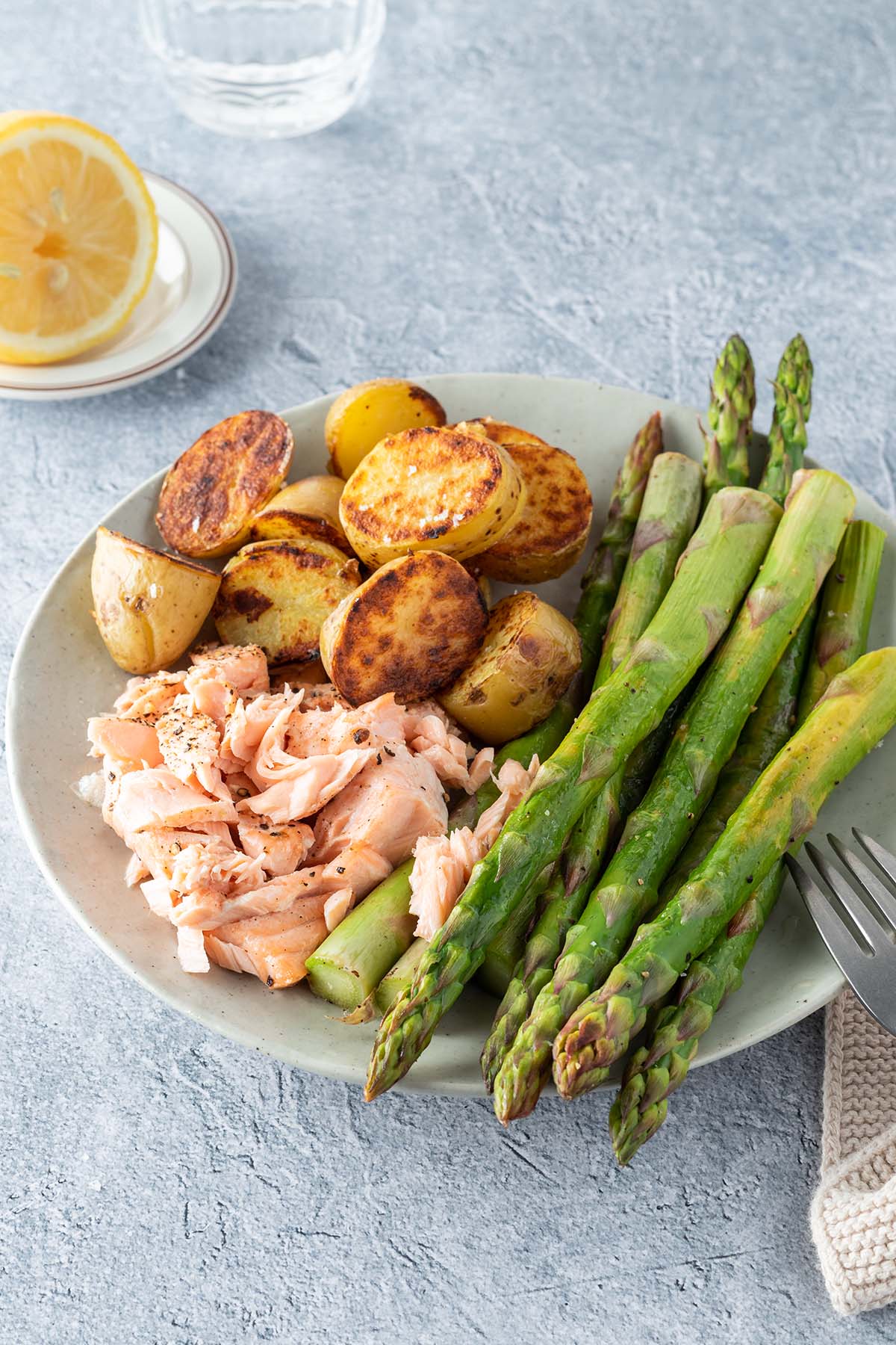 Ein Teller mit gr&uuml;nem Spargel, knusprig angebratenen Kartoffeln und ein kleine St&uuml;cke zerrissener Lachs, eine Glas Wasser und eine Zitronenh&auml;lfte im Hintergrund.