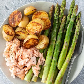 Gr&uuml;ner Spargel mit zerrupftem Lachs und knusprig angebratenen Kartoffelh&auml;lften auf einem Teller.