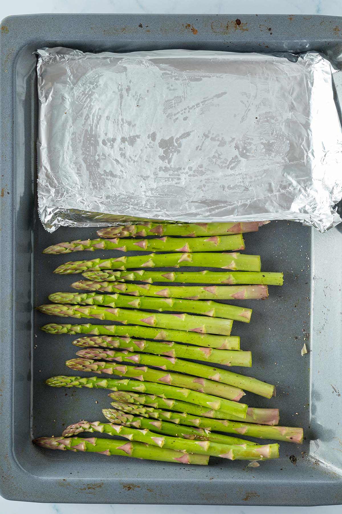 Vogelperspektive auf ein Backblech mit gr&uuml;nem Spargel und einem St&uuml;ck Aluminiumfolie daneben.