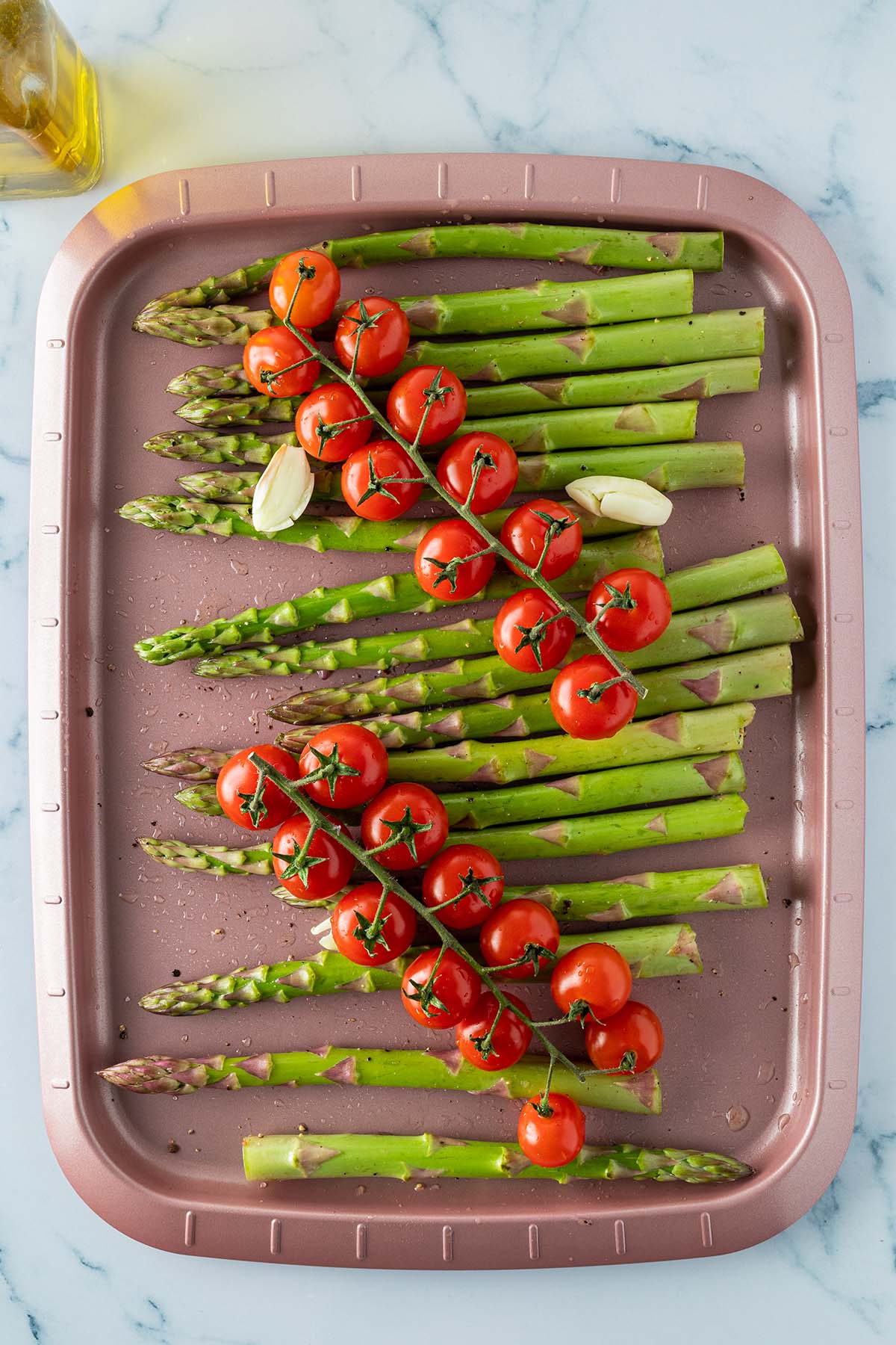 Gr&uuml;ne Spargelstangen mit gesch&auml;lten Knoblauchzehen und zwei Tomatenrispen auf einem Backblech.