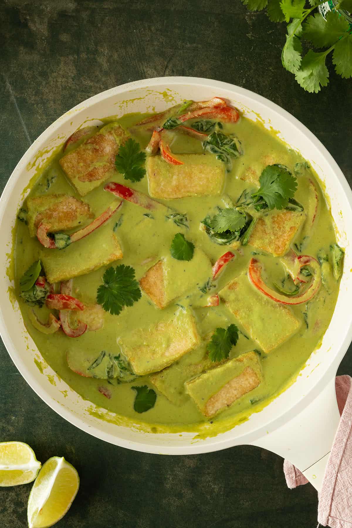 Cremiges gr&uuml;nes Thai Curry serviert in einer Pfanne mit Gem&uuml;se und knusprigen Tofu-Scheiben auf einem dunkelgr&uuml;nen Untergrund.