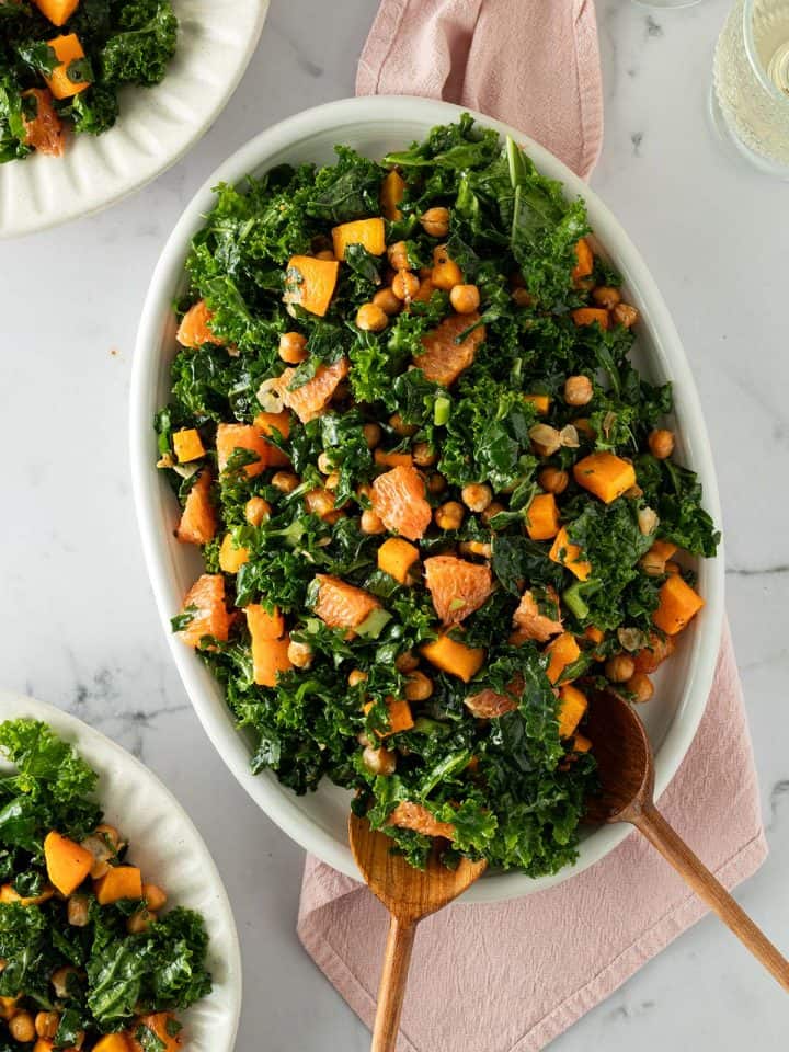 Wintersalat aus Gr&uuml;nkohl, Orange, K&uuml;rbis und Kichererbsen serviert auf einem ovalen Teller mit Salatbesteck. Zwei Teller mit Salat und Weingl&auml;ser im Bildrand.