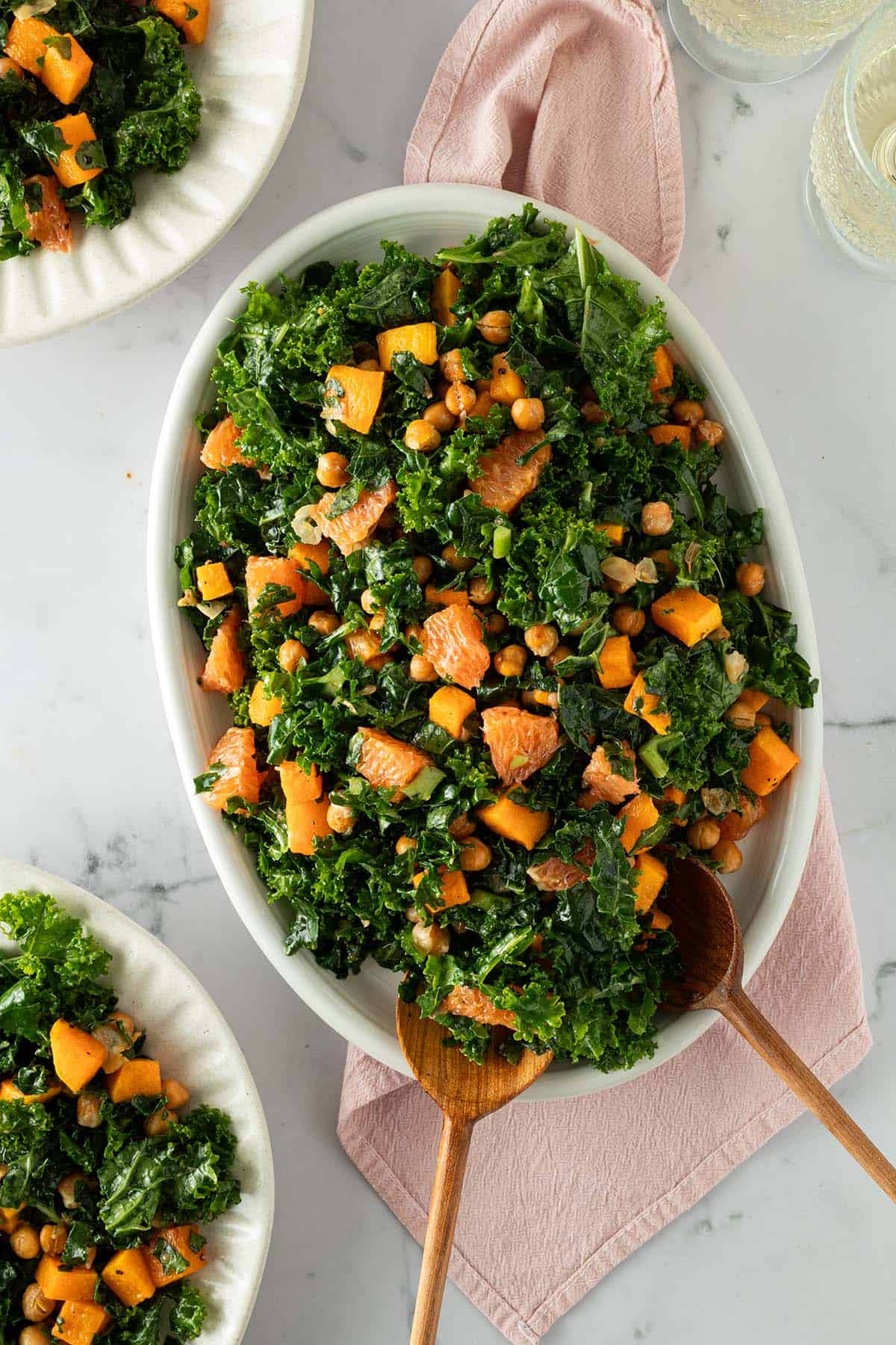 Wintersalat aus Gr&uuml;nkohl, Orange, K&uuml;rbis und Kichererbsen serviert auf einem ovalen Teller mit Salatbesteck. Zwei Teller mit Salat und Weingl&auml;ser im Bildrand.