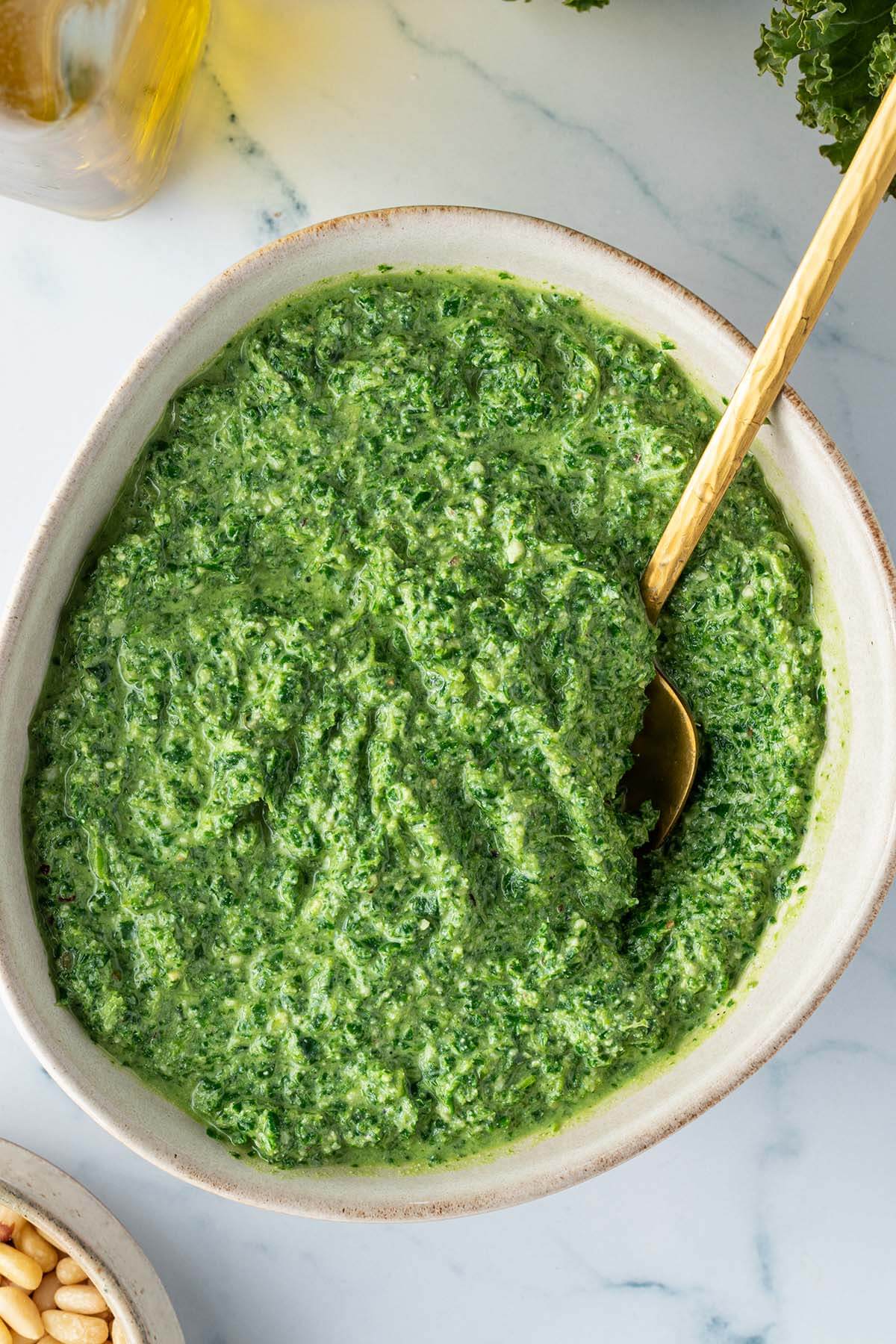 Sch&uuml;ssel mit einer Gr&uuml;nkohl Pesto auf einem Marmor-Untergrund, mit einer Flasche &Ouml;l und einer Sch&uuml;ssel Pinienkerne im Bildrand.