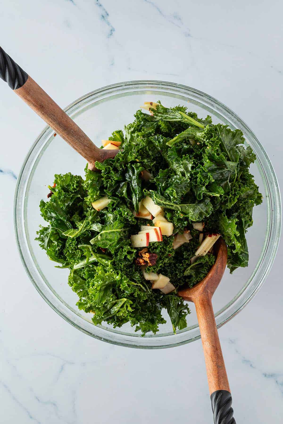 Massierter Gr&uuml;nkohl wird mit den Apfelst&uuml;cken und ger&ouml;steten N&uuml;ssen in einer Sch&uuml;ssel mit Salatbesteck vermischt.