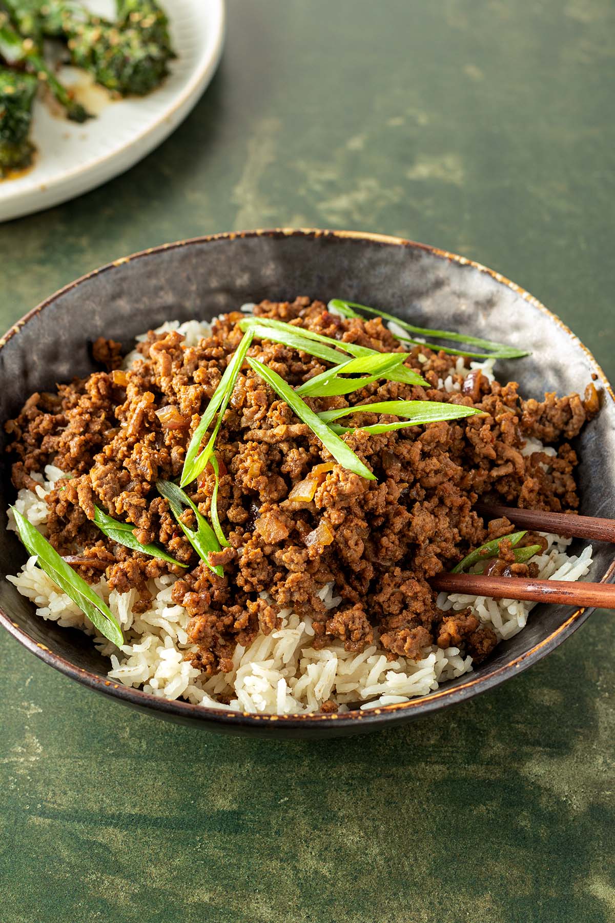 Eine Sch&uuml;ssel mit Reis, asiatischem Hackfleisch und Fr&uuml;hlingszwiebeln, im Hintergrund ein Teller mit Brokkoli.