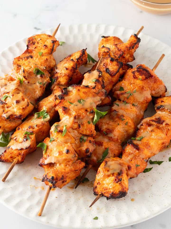 Mehrere Chicken Satay H&auml;hnchenspie&szlig;e auf einem Teller, serviert mit einer Erdnusssauce und frischen Kr&auml;utern.