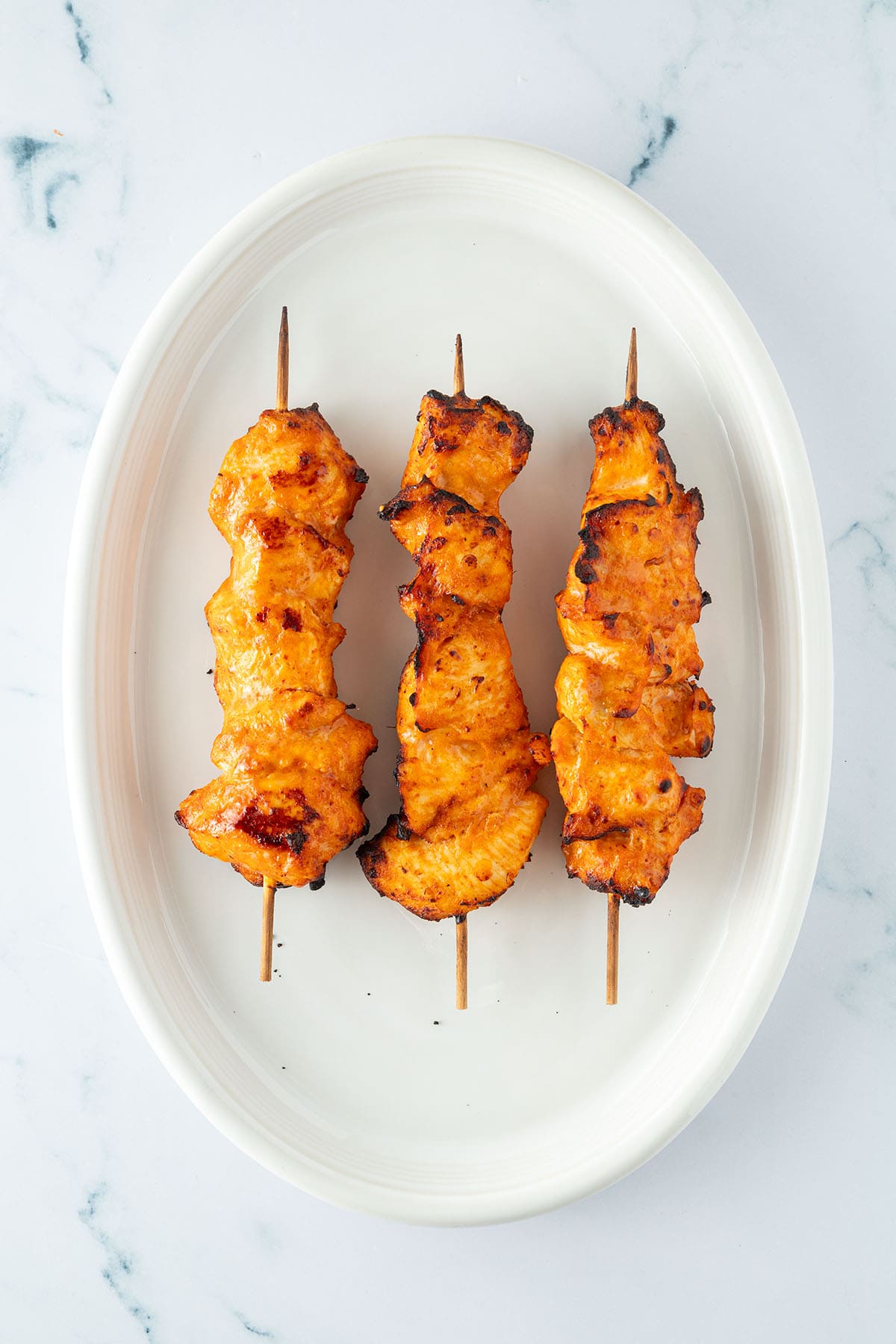 Drei fertig gegarte Chicken Satay Spie&szlig;e auf einem ovalen Teller.