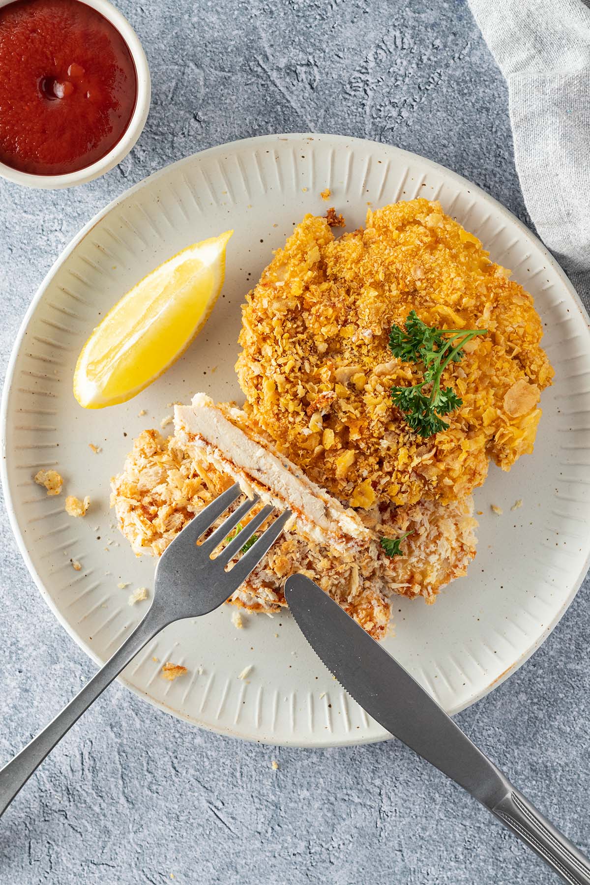 Teller mit zwei H&auml;hnchenschnitzeln, wo mit Gabel und Messer ein Bissen abschnitten wurde. Sch&uuml;ssel Ketchup und eine Serviette im Bildrand.
