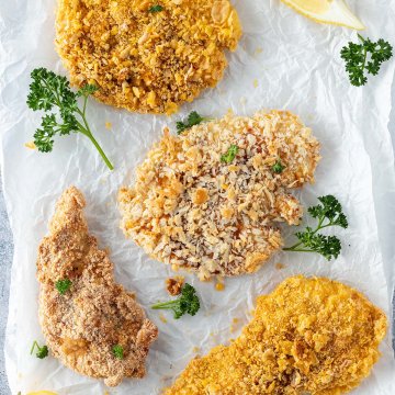 Vier unterschiedlich panierte H&auml;hnchenschnitzel serviert mit frischer Petersilie und Zitrone.