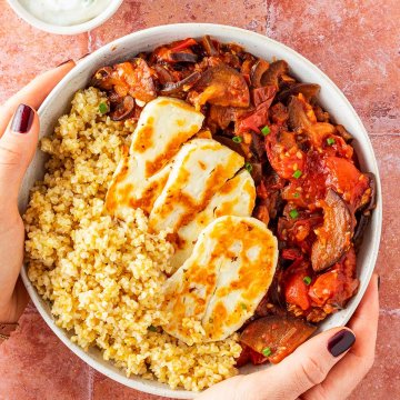 Zwei H&auml;nde halten eine Bowl mit Halloumi, Bulgur und einem Tomatengem&uuml;se vor einem roten Fliesenuntergrund.