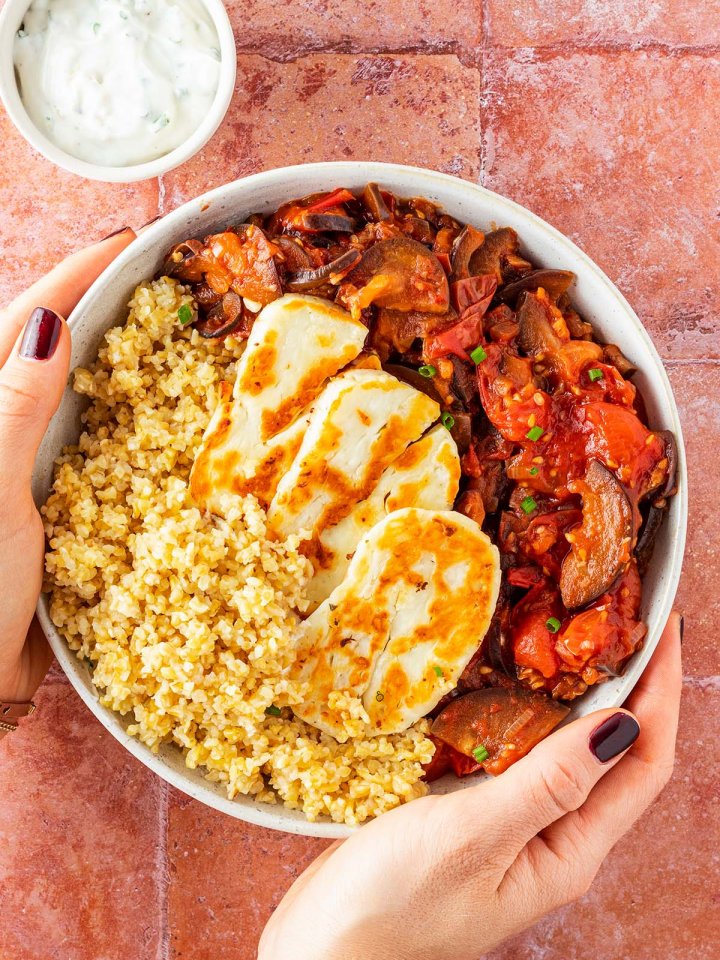 Zwei H&auml;nde halten eine Bowl mit Halloumi, Bulgur und einem Tomatengem&uuml;se vor einem roten Fliesenuntergrund.