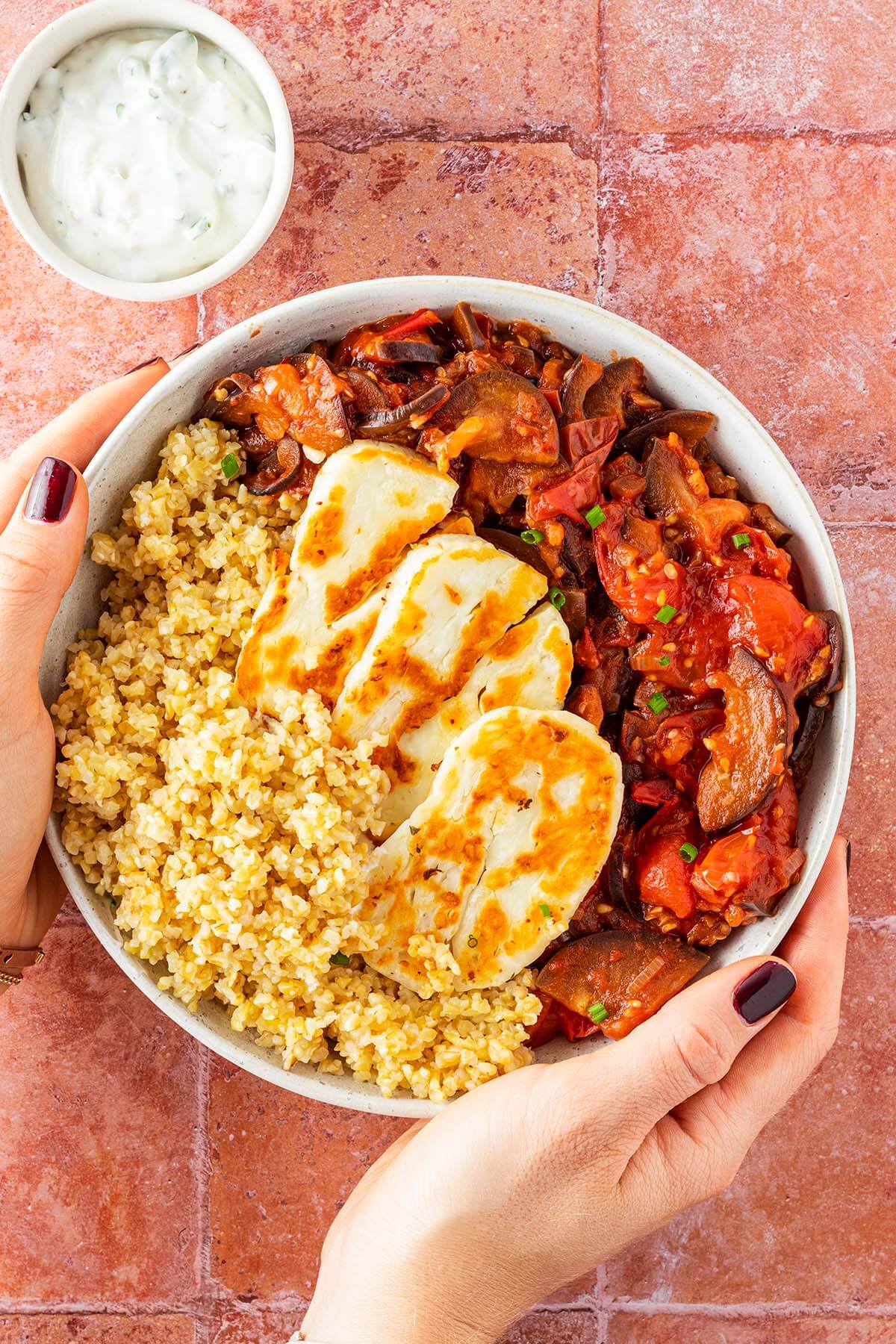 Zwei H&auml;nde halten eine Bowl mit Halloumi, Bulgur und einem Tomatengem&uuml;se vor einem roten Fliesenuntergrund.