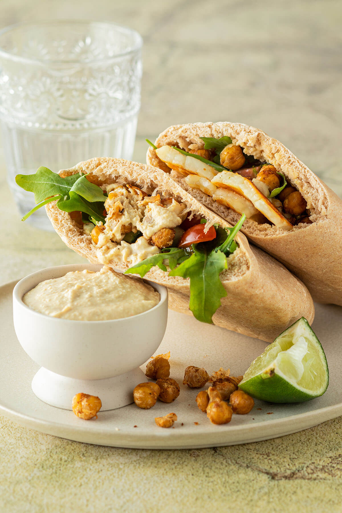 Zwei mit Halloumi, Kichererbsen, Gem&uuml;se und Hummus gef&uuml;llte Pita Brote auf einem Teller mit einer Sch&uuml;ssel Hummus, ein Glas mit Wasser im Hintergrund.