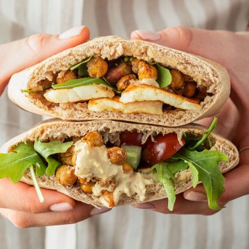 Eine Person mit Sch&uuml;rze h&auml;lt zwei mit Halloumi, Kichererbsen und Hummus gef&uuml;llte Pita Taschen in der Hand.