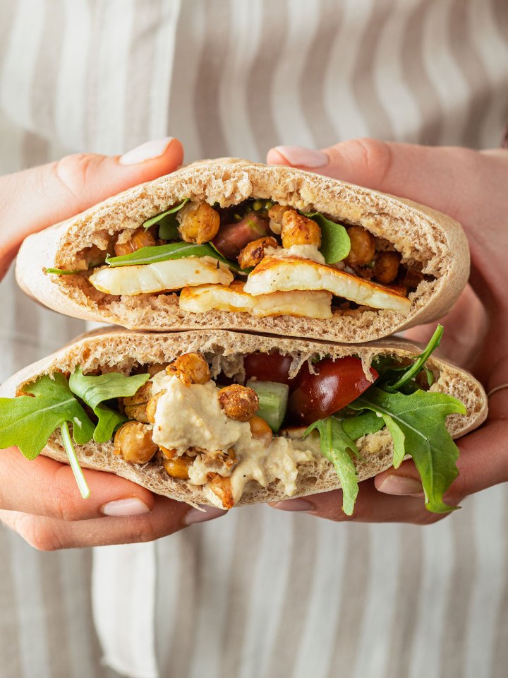 Eine Person mit Sch&uuml;rze h&auml;lt zwei mit Halloumi, Kichererbsen und Hummus gef&uuml;llte Pita Taschen in der Hand.