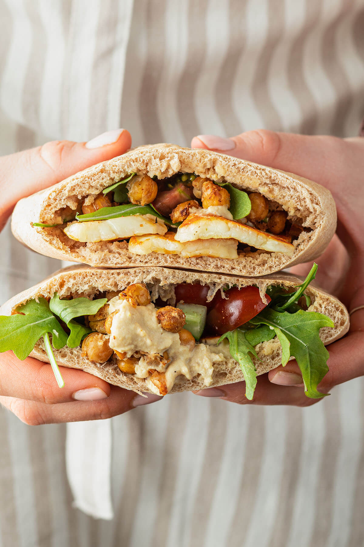 Eine Person mit Sch&uuml;rze h&auml;lt zwei mit Halloumi, Kichererbsen und Hummus gef&uuml;llte Pita Taschen in der Hand.