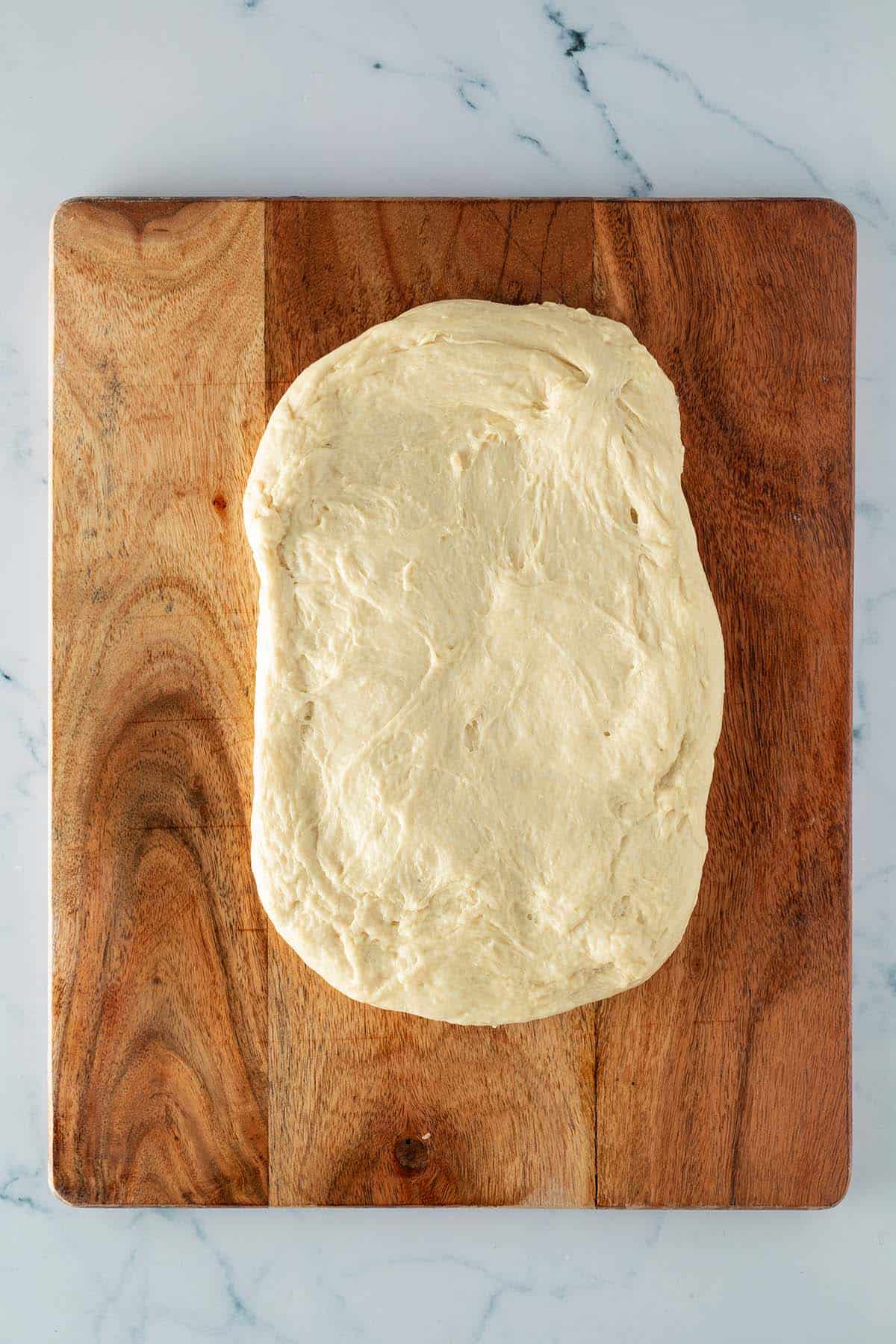 Zu einem l&auml;nglichen Rechteck flachgedr&uuml;ckter Hefeteig auf einem Holzbrett.
