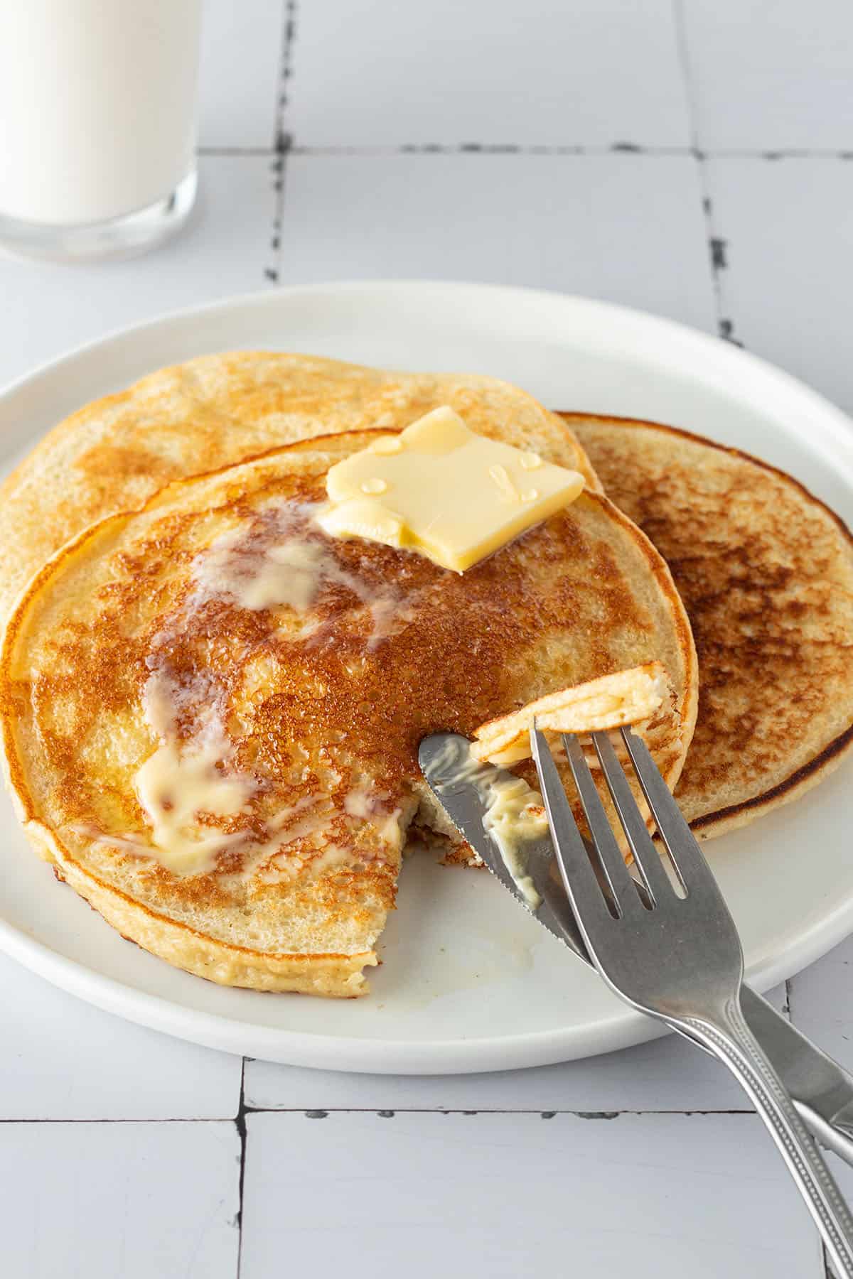 Ein Teller mit drei High Protein H&uuml;ttenk&auml;se Pancakes auf einem wei&szlig;en Flie&szlig;en-Untergrund und einem Glas Milch im Hintergrund.