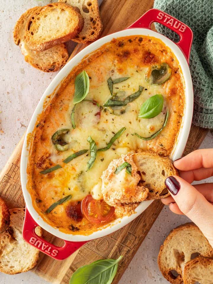 Eine Hand tunkt einen Brotchip in den warmen, mit K&auml;se &uuml;berbackenen H&uuml;ttenk&auml;se Dip ein.