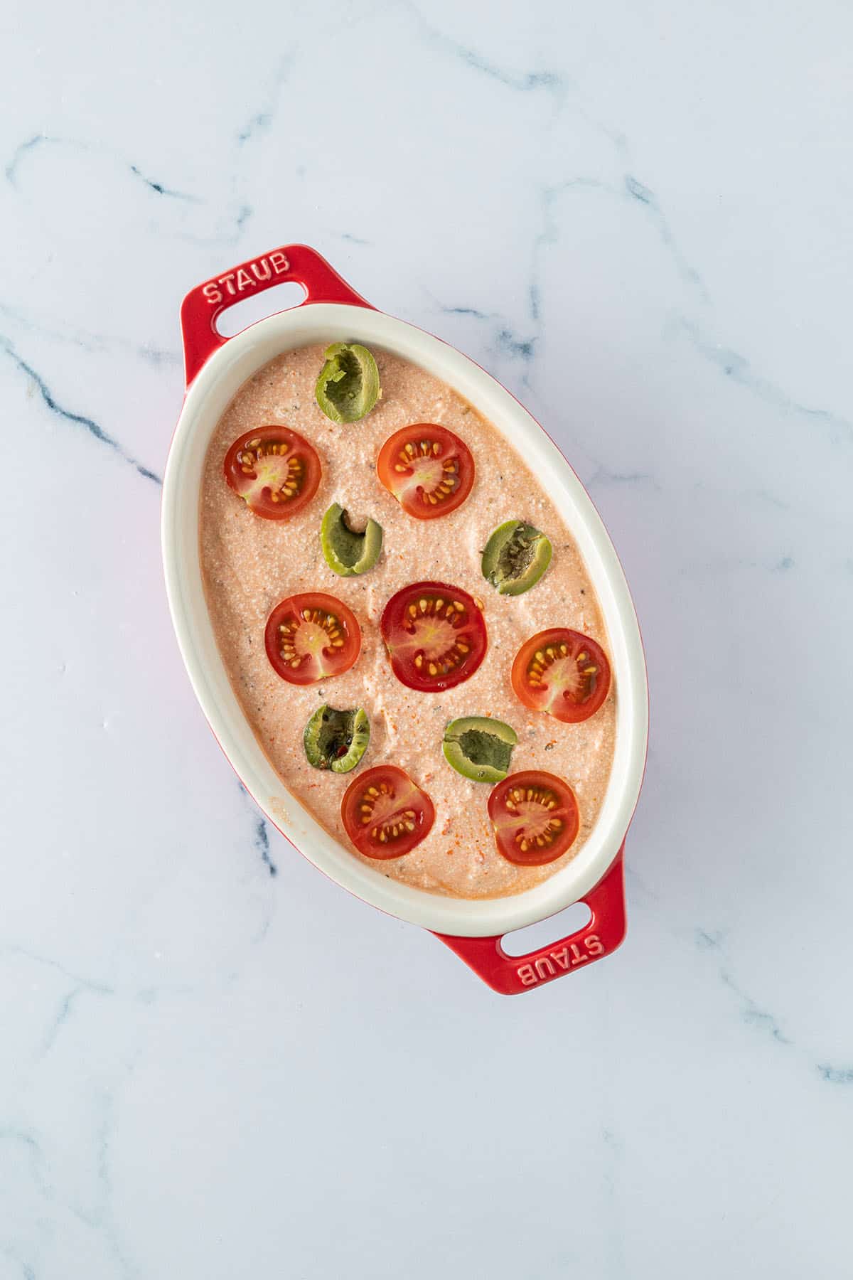 H&uuml;ttenk&auml;se-Tomatencreme in einer kleinen Auflaufform mit halbierten Kirschtomaten und Oliven auf der Oberfl&auml;che.