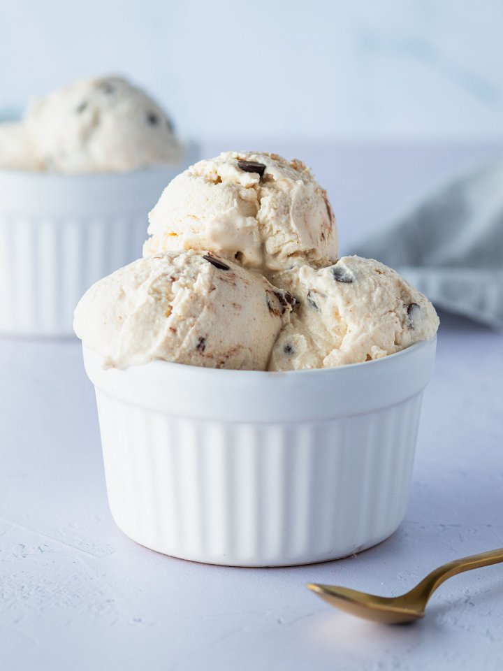 Mehrere Cottage-Cheese-Eis-Kugeln mit Schokost&uuml;ckchen in einer wei&szlig;en Sch&uuml;ssel, im Hintergrund eine zweite Sch&uuml;ssel mit Eiskugeln.
