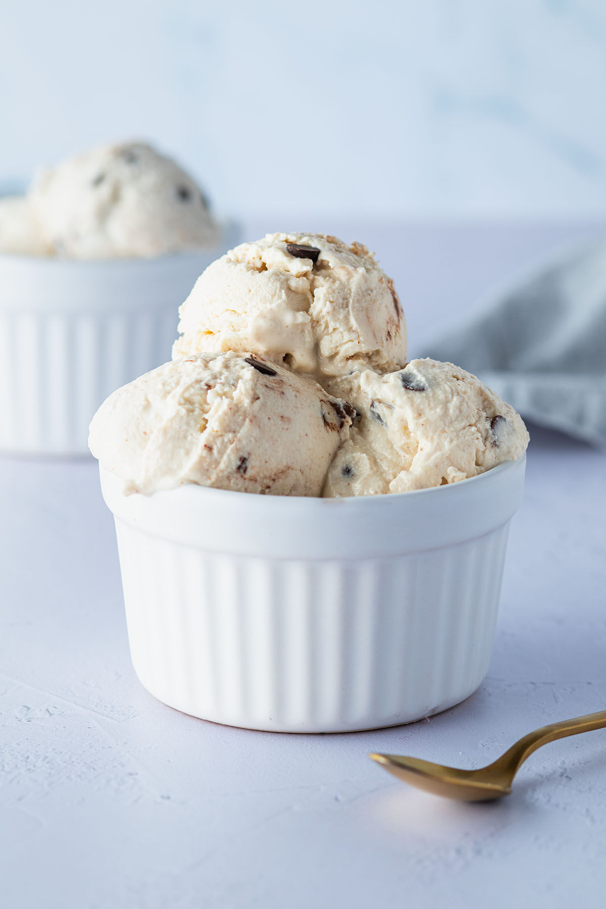 Mehrere Cottage-Cheese-Eis-Kugeln mit Schokost&uuml;ckchen in einer wei&szlig;en Sch&uuml;ssel, im Hintergrund eine zweite Sch&uuml;ssel mit Eiskugeln.