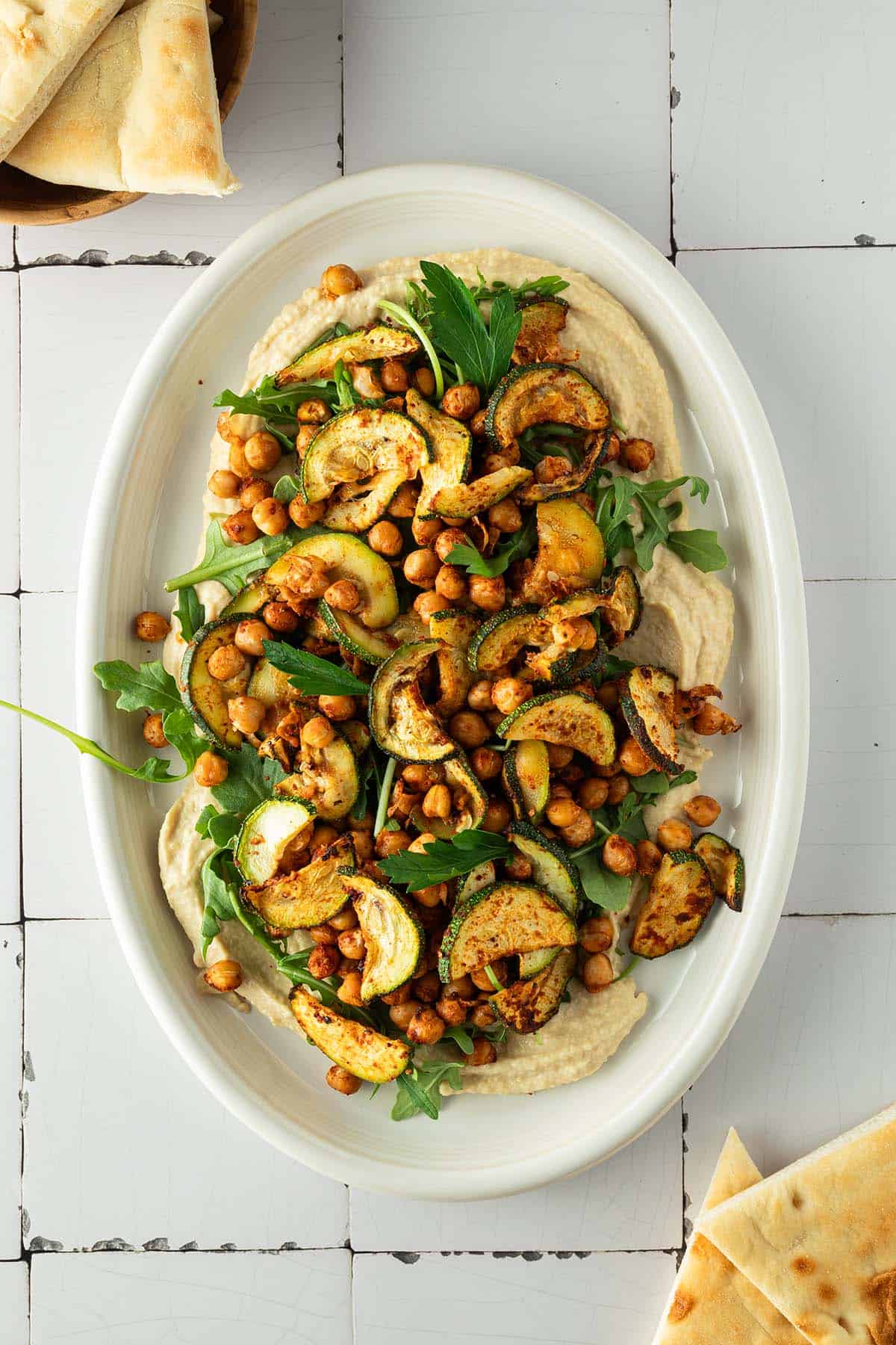 Hummus Platte mit ger&ouml;steten Kichererbsen, Zucchini und Rucola, Pita-Dreieche im Bildrand.