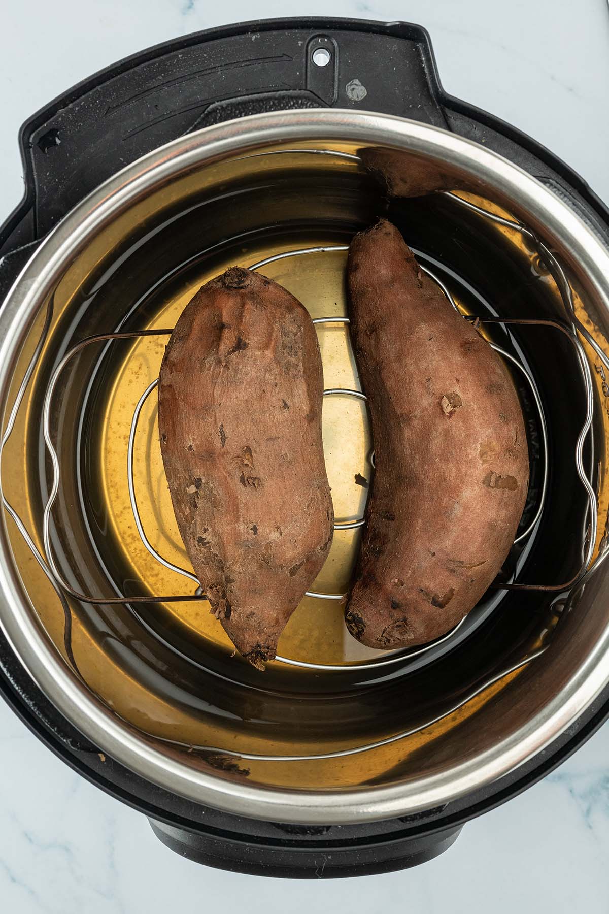 Vogelperspektive in den offenen Instant Pot, wo zwei gekochte S&uuml;&szlig;kartoffeln auf einem Metallgestell liegen.