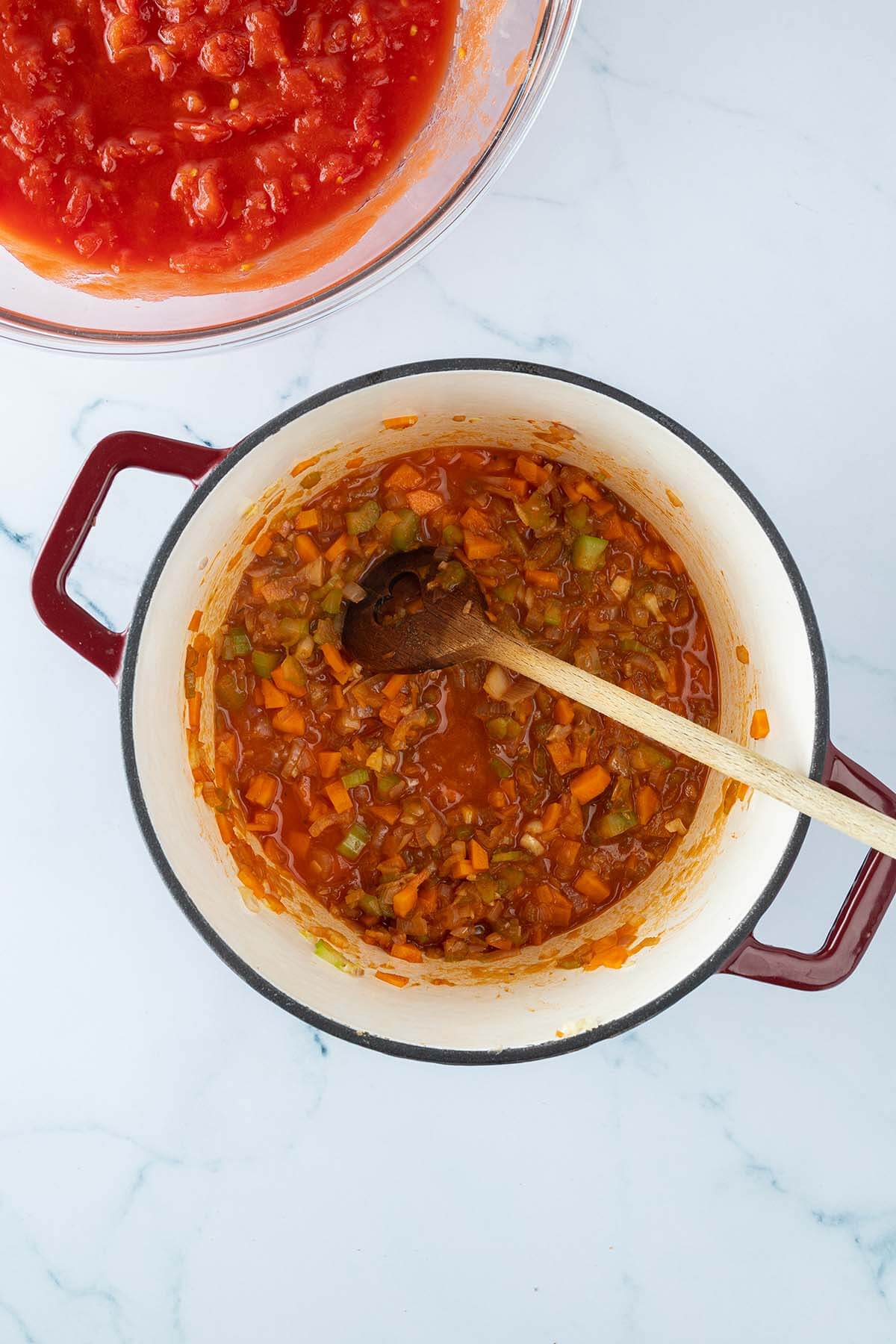 Vogelperspektive auf einen Topf mit angebratenem Gem&uuml;se, das mit etwas Fl&uuml;ssigkeit abgel&ouml;scht wurden. Daneben eine Glassch&uuml;ssel mit st&uuml;ckigen Tomaten.