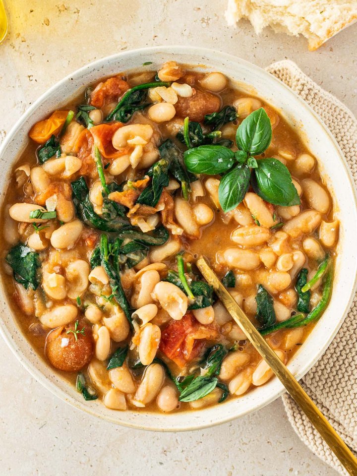 Teller mit einer mediterranen wei&szlig;en Bohnenpfanne in Tomatensauce mit Spinat, serviert mit Brot und frischem Basilikum.