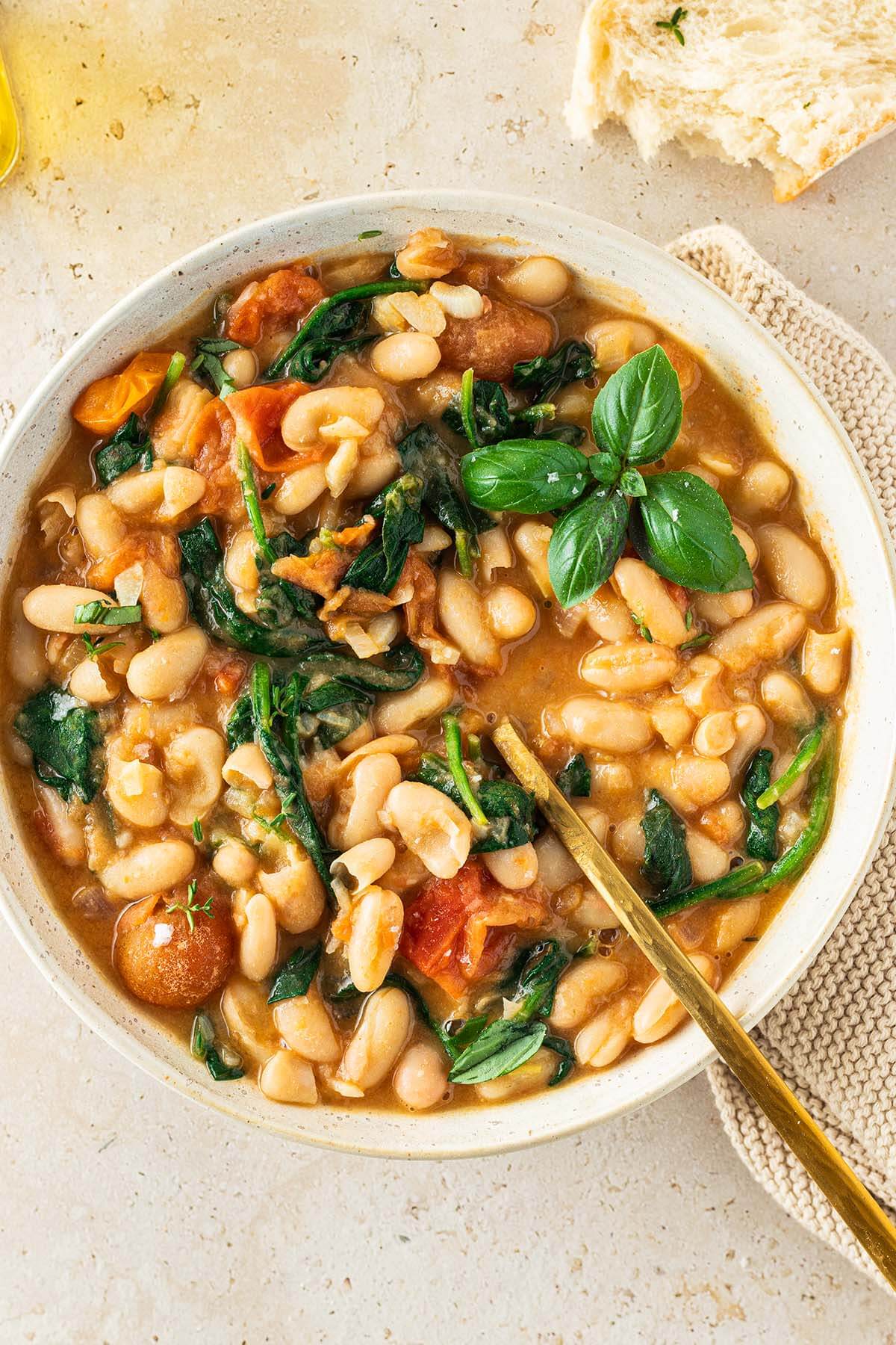 Teller mit einer mediterranen wei&szlig;en Bohnenpfanne in Tomatensauce mit Spinat, serviert mit Brot und frischem Basilikum.