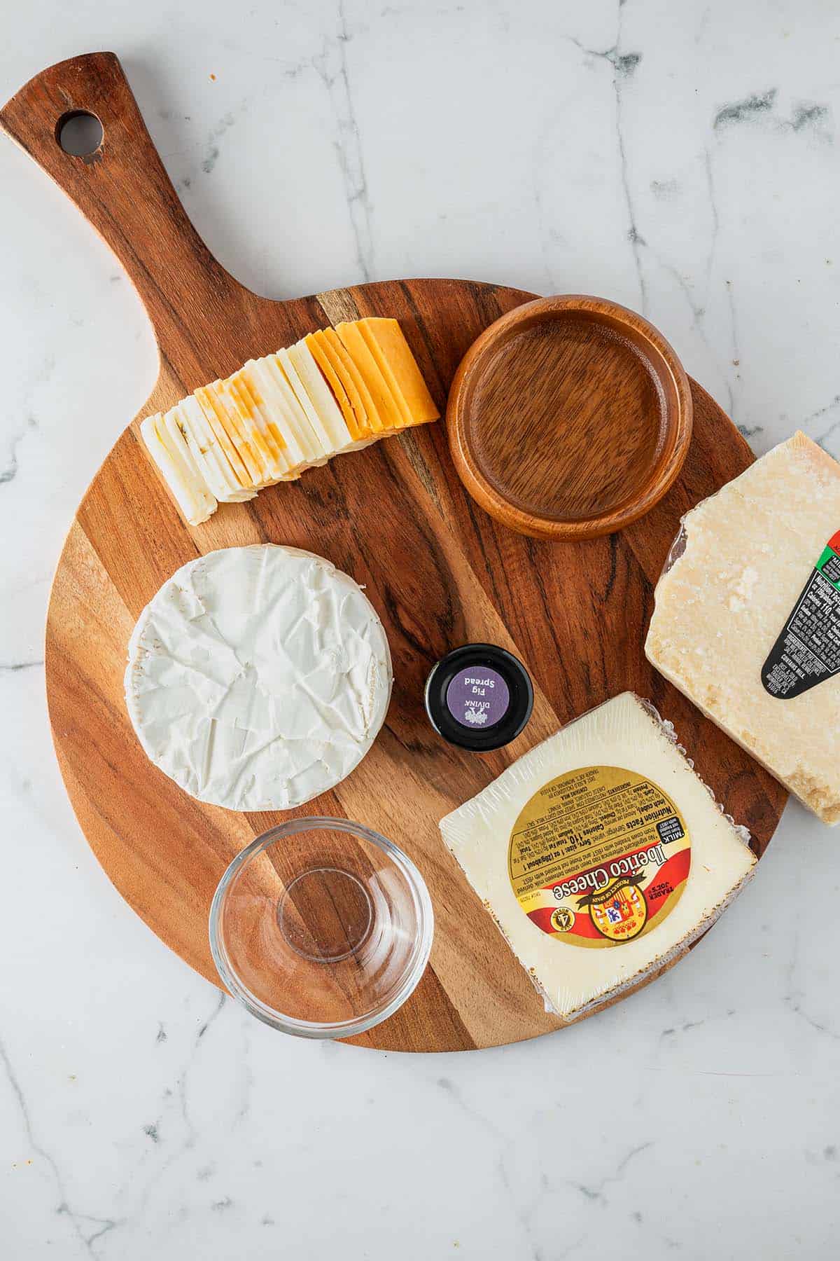 Holzbrett mit noch verpacktem K&auml;se, Sch&auml;lchen und vorbereitetem Zubeh&ouml;r f&uuml;r eine K&auml;seplatte.